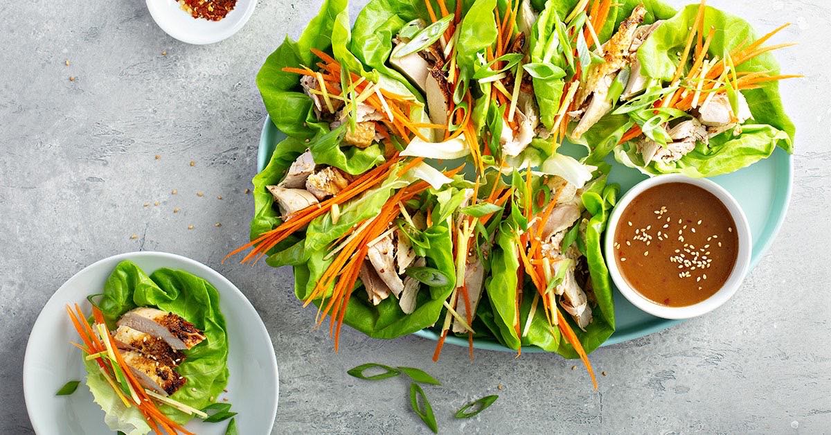 Plate of chicken lettuce wraps with carrots, scallions, and dipping sauce arranged neatly on a light table.