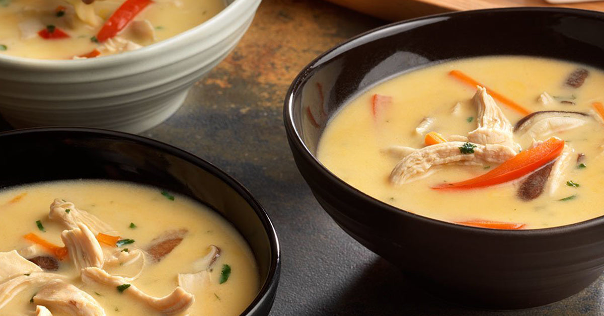 Two bowls of creamy chicken soup are shown, one black and one light gray, filled with shredded chicken, colorful vegetables, and herbs, set against a textured dark surface.