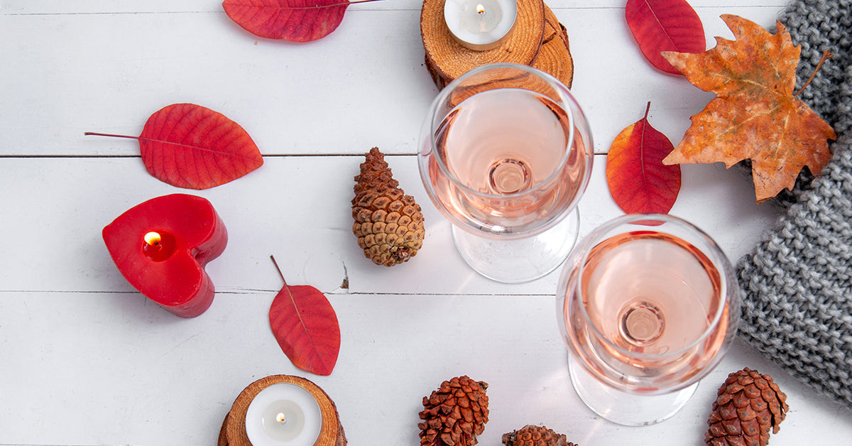 A cozy autumn flat lay on white wood featuring two glasses of rosé wine, scattered red leaves, pinecones, a lit red heart-shaped candle, and a grey knitted blanket, evoking a quiet, intimate holiday celebration.