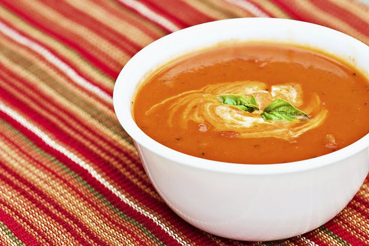Bowl of creamy tomato soup with a swirl of cream and basil leaves on top, set on a red and green striped cloth.
