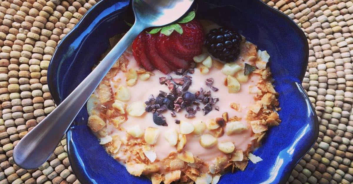 Blue bowl of yogurt topped with granola, cacao nibs, sliced strawberries, and a blackberry with a spoon on top.