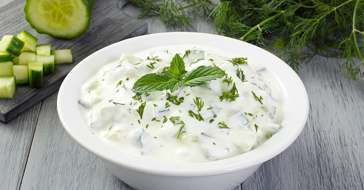 A bowl of creamy cucumber yogurt sauce garnished with fresh mint and herbs sits on a wooden surface, with chopped cucumbers nearby and greenery in the background.
