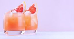 Two grapefruit cocktails with ice and citrus slices in clear glasses against a soft pink background.