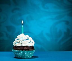 Chocolate cupcake with white frosting and a lit blue candle against a blue background.