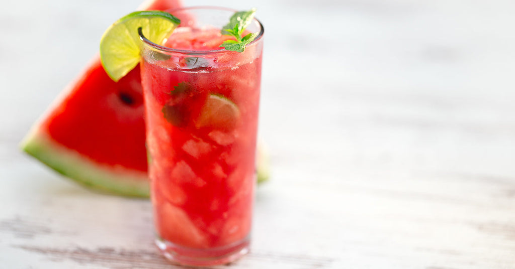 Tall glass of watermelon lime drink with ice and mint, served beside a fresh watermelon slice.