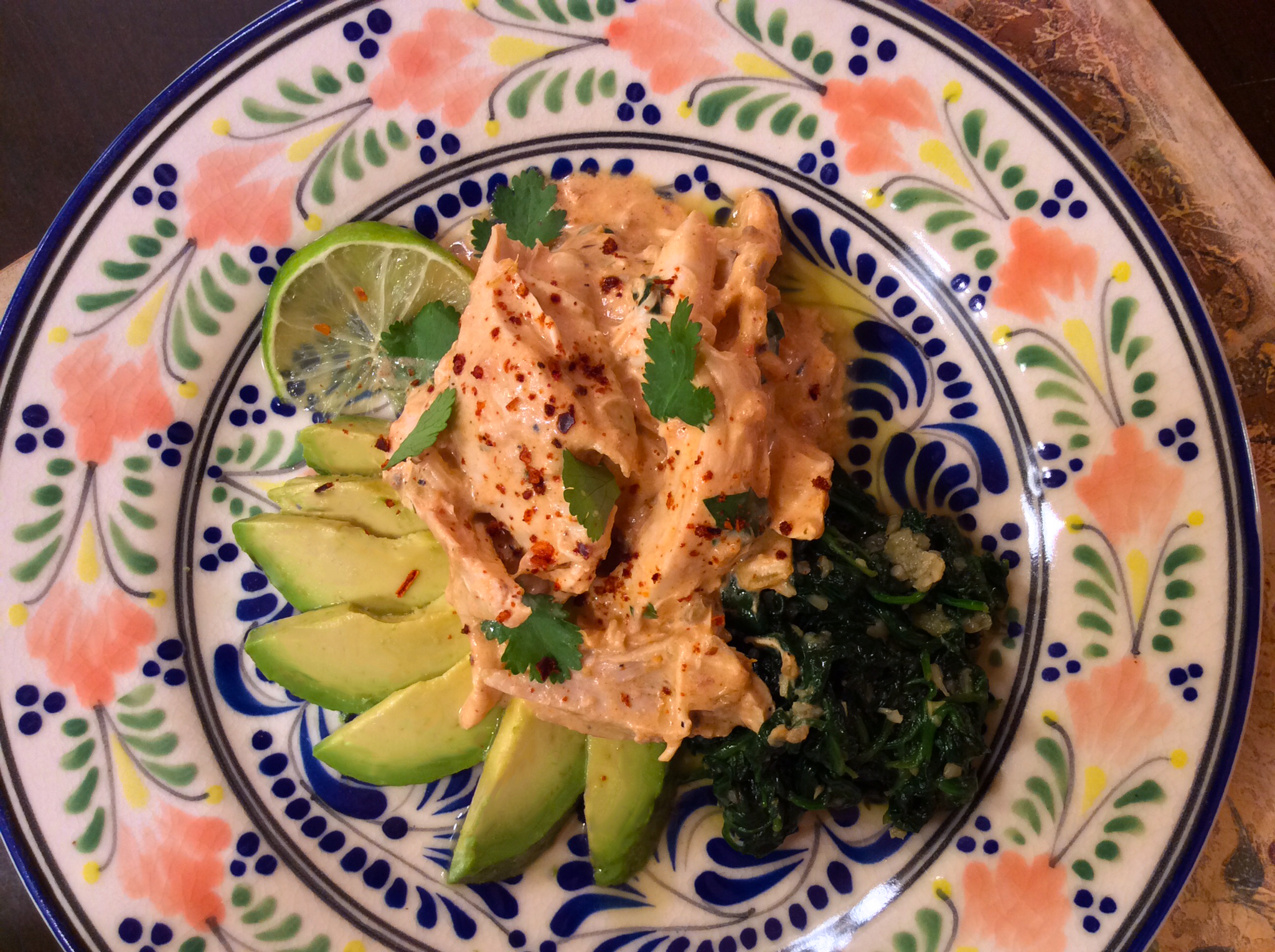 Creamy spiced chicken served with avocado slices, sautéed greens, lime, and cilantro on a colorful patterned plate.