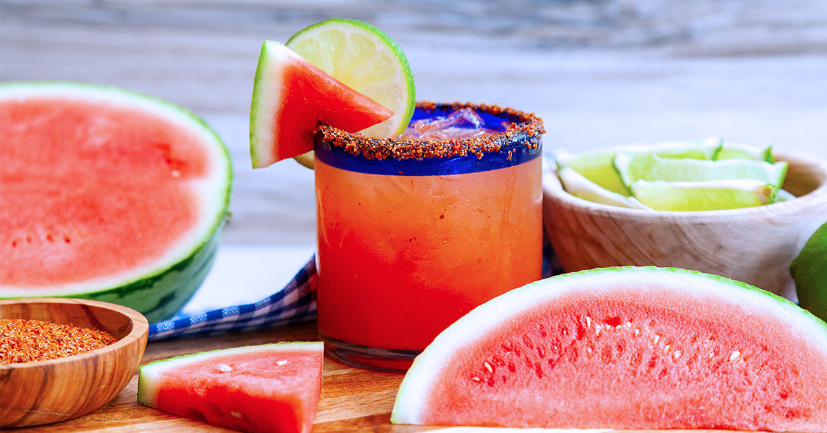 A vibrant watermelon margarita sits on a wooden surface, garnished with a lime wedge and a watermelon slice, surrounded by fresh watermelon pieces and a bowl of lime wedges.
