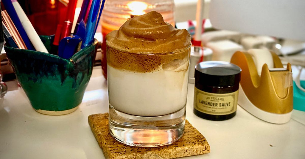 Glass of whipped dalgona coffee on a coaster, sitting on a desk with pens, a candle, and a jar of lavender salve.