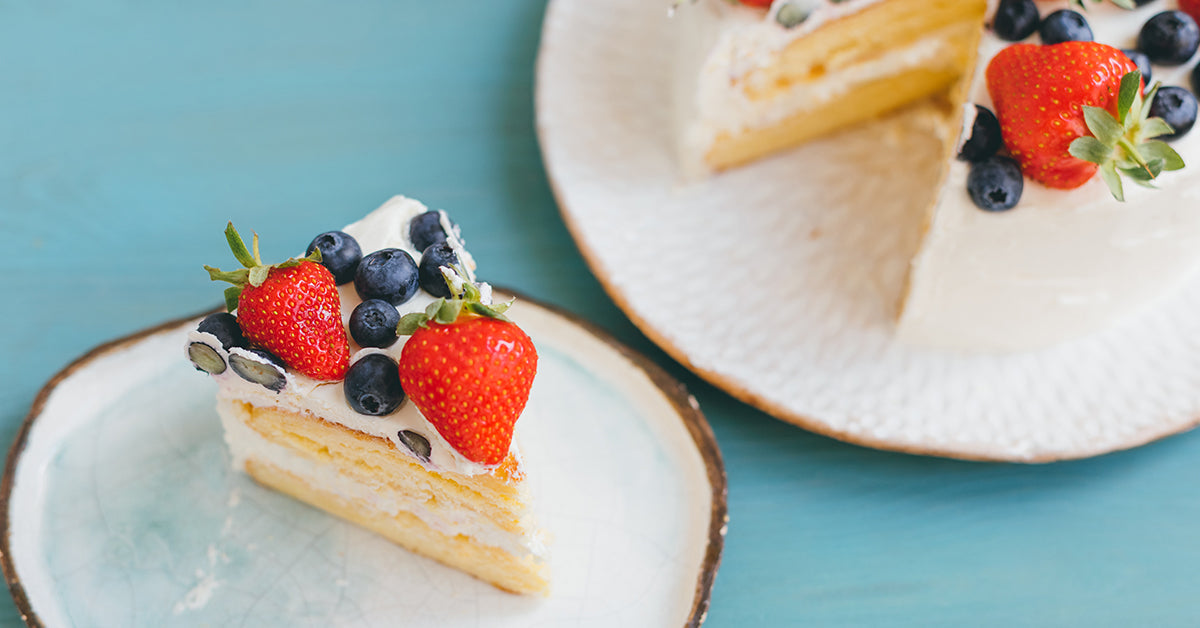 A slice of whipped cream cake topped with fresh strawberries and blueberries sits on a small plate, next to a larger cake on a textured white plate, all set against a light blue background.