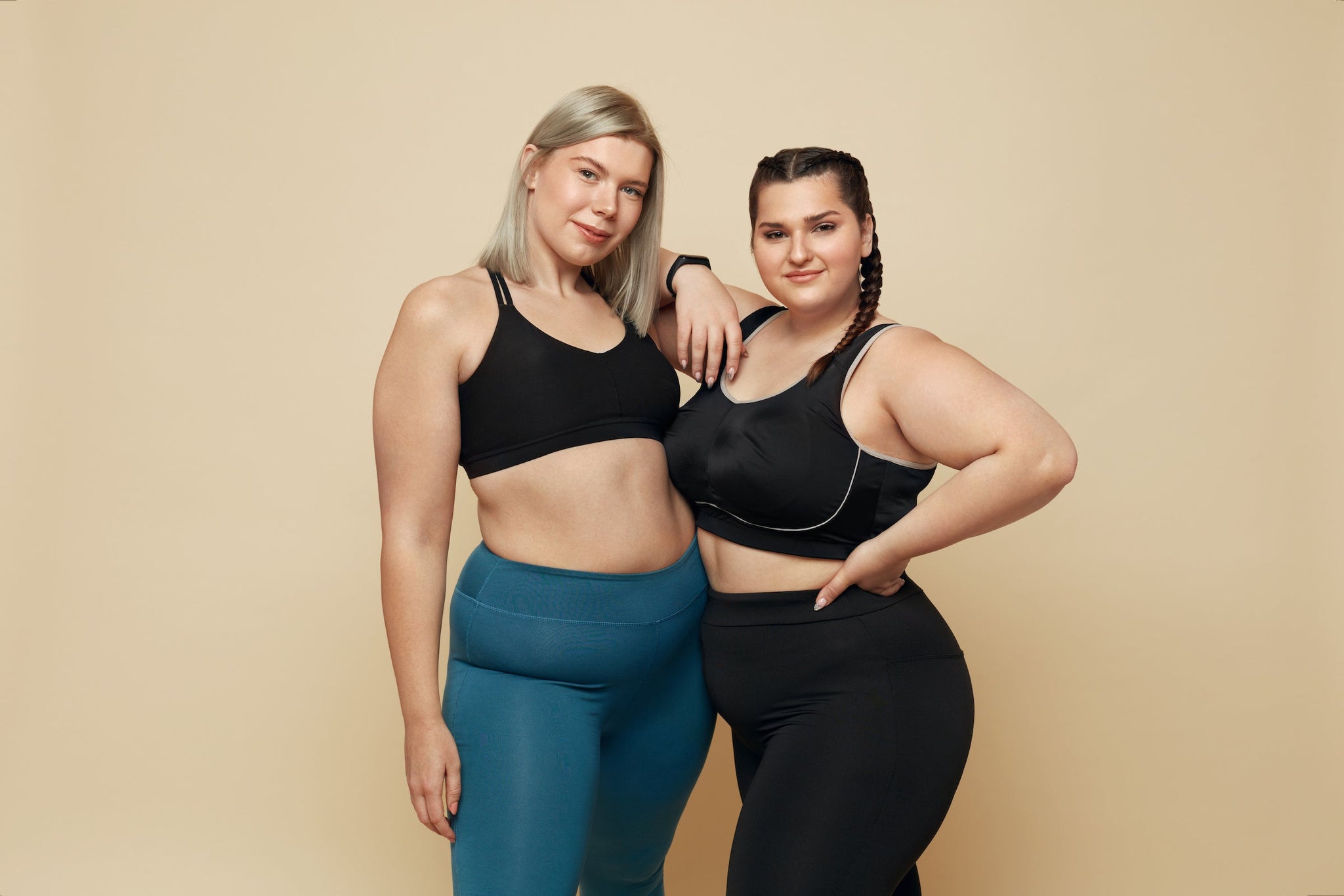 Two women in athletic wear posing confidently together against a plain background, smiling at the camera
