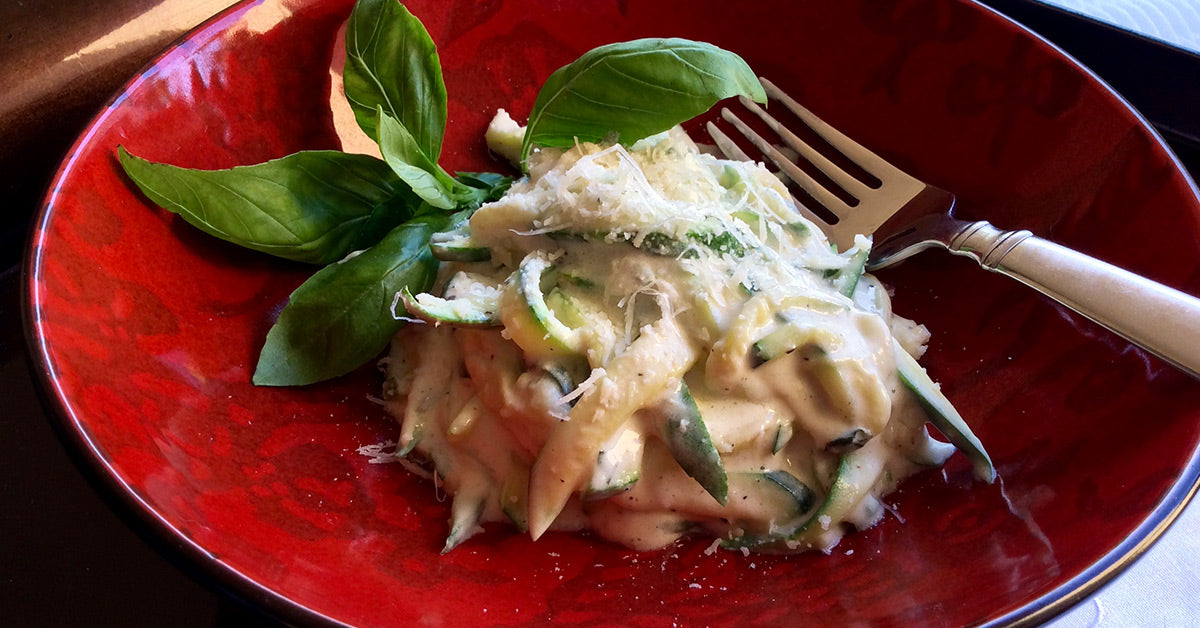 Creamy zucchini noodle dish topped with grated cheese and basil on a red plate with a fork.