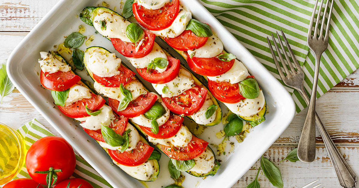 Three zucchini halves are layered with slices of tomato and mozzarella cheese, garnished with fresh basil, and arranged in a white dish on a striped green and white tablecloth, with tomatoes and olive oil nearby.