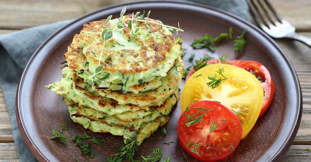 Stack of zucchini fritters on a plate with sliced red and yellow tomatoes and fresh herbs.