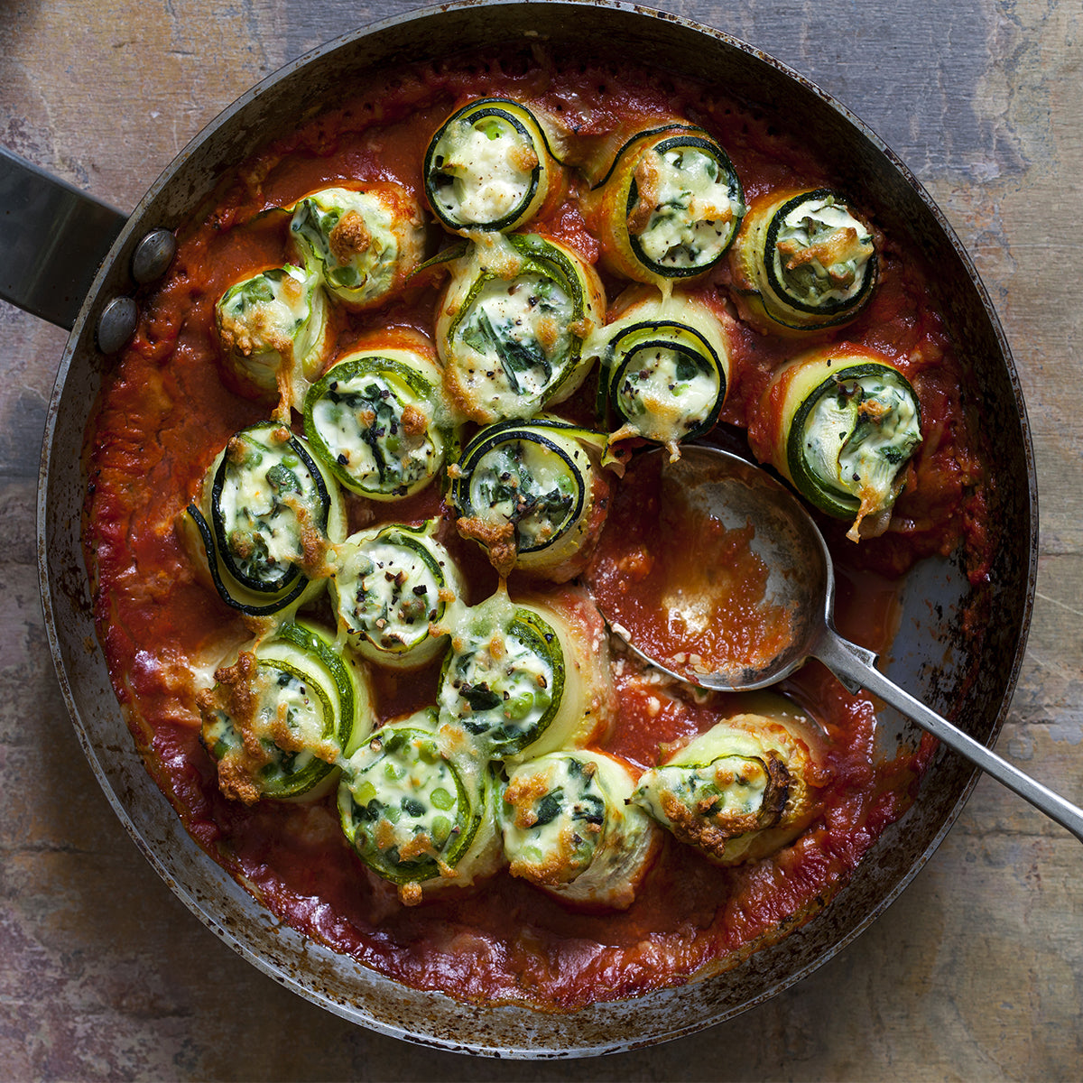 Zucchini rolls filled with ricotta and spinach are baked in a rich tomato sauce, displayed in a metal pan with a serving spoon resting beside them.