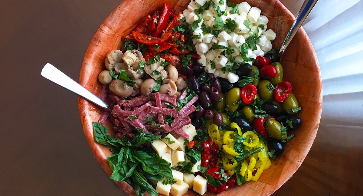 Colorful antipasto salad in a wooden bowl with cheeses, olives, salami, peppers, mushrooms, tomatoes, and herbs.