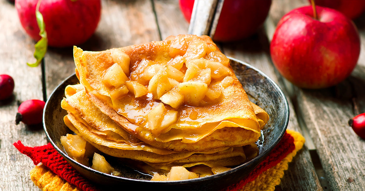 A stack of golden crepes topped with warm, caramelized apple chunks sits in a black bowl on a rustic wooden table, surrounded by fresh red apples.