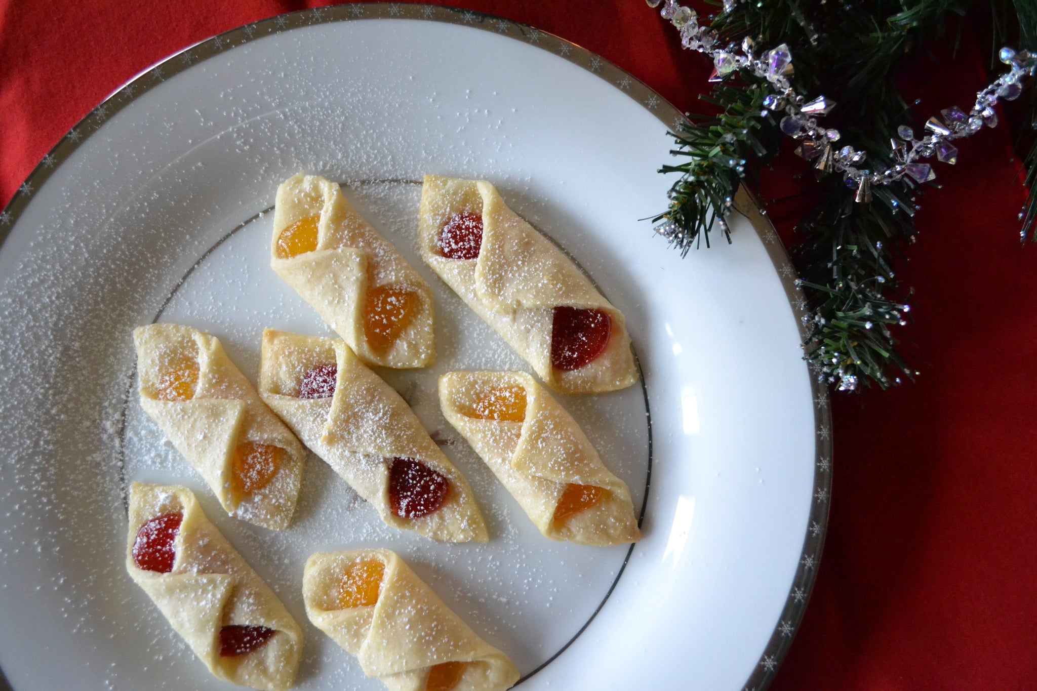 Plate of powdered sugar-dusted cookies filled with red and orange jam, set on a festive red tablecloth with greenery.