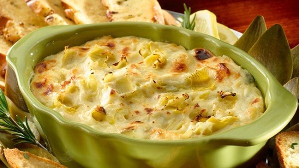 Creamy baked artichoke dip in a green dish, lightly browned on top and served with toasted bread slices.