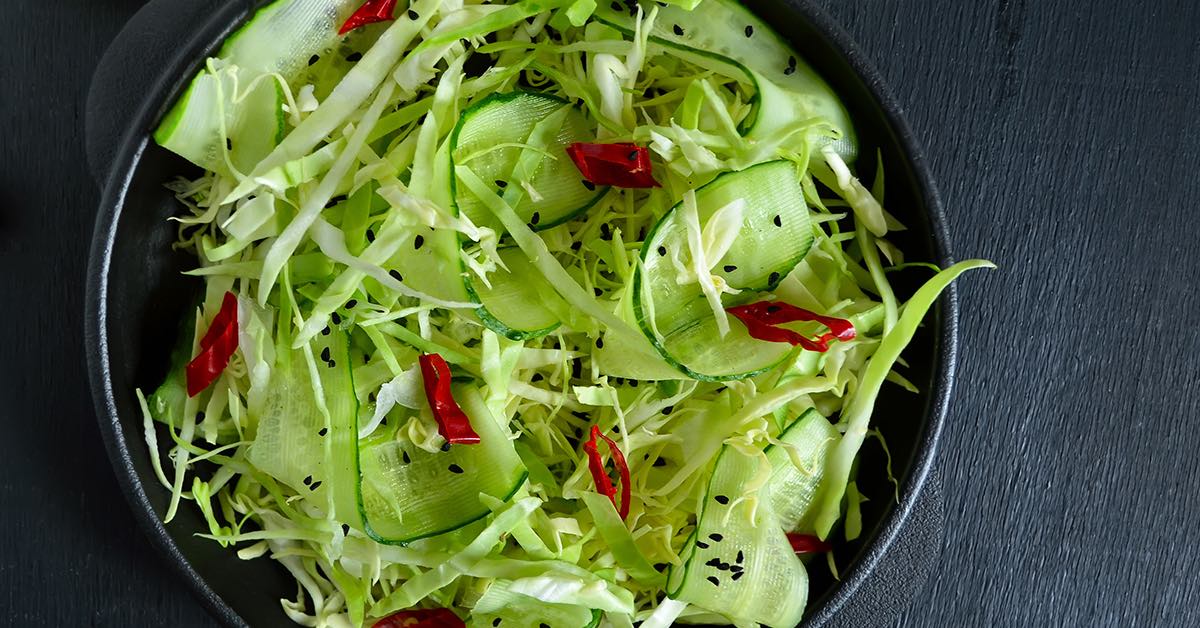 Shredded cabbage and ribboned cucumber salad with red chili slices and black sesame seeds in a dark bowl.
