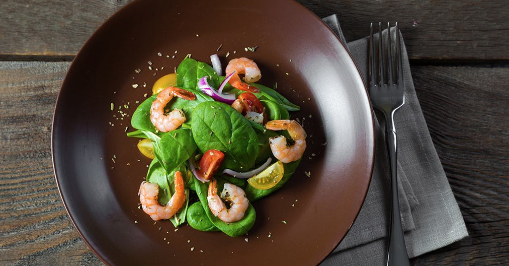 Shrimp and spinach salad with cherry tomatoes and red onion on a brown plate beside a fork on a napkin.
