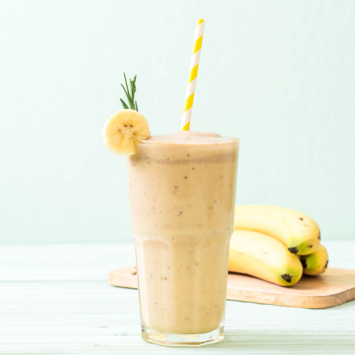 A tall glass of banana smoothie with a yellow straw and a banana slice garnish sits on a light wooden surface, with a bunch of ripe bananas in the background against a pale green wall.