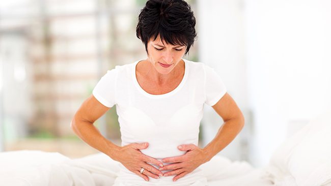 Woman sitting on a bed holding her stomach with a pained expression.