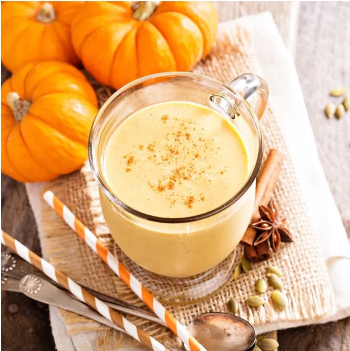 A glass of creamy pumpkin smoothie topped with a sprinkle of cinnamon sits on a burlap cloth, surrounded by small pumpkins, a spoon, and striped straws, evoking a cozy autumn vibe.