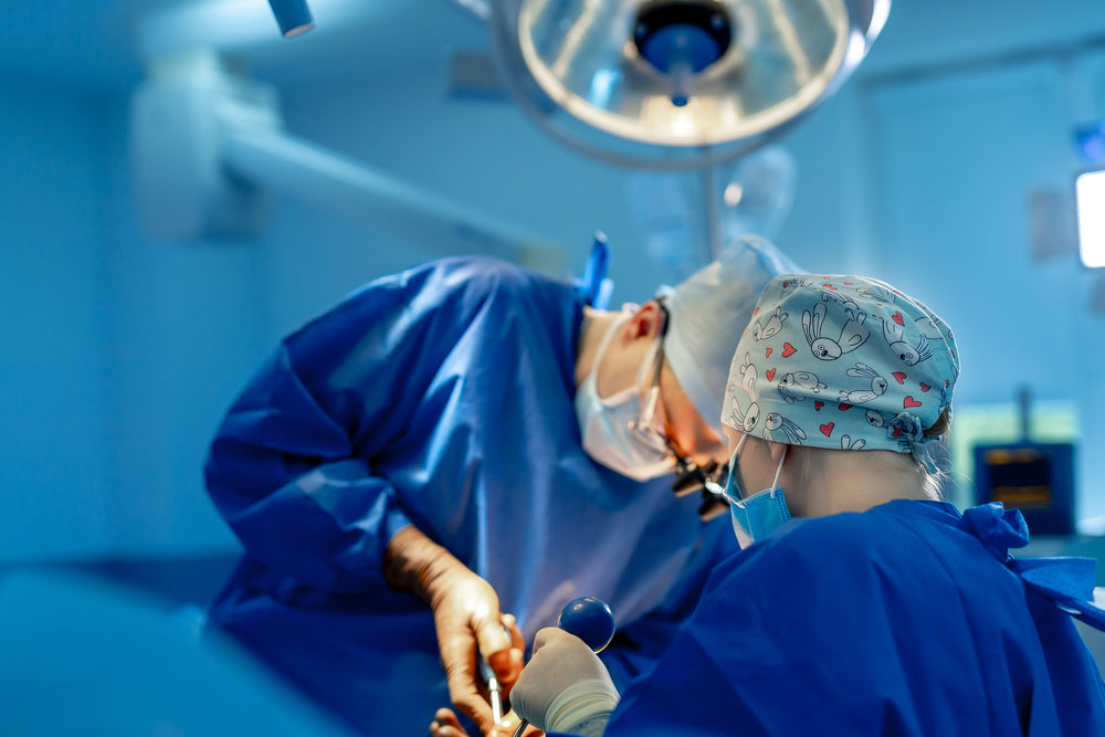 Two medical professionals in blue scrubs and masks are focused on a surgical procedure, with one holding instruments and the other assisting, in a bright, sterile operating room.