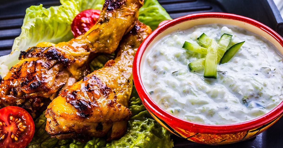 Grilled chicken drumsticks on lettuce served with a bowl of creamy cucumber dip topped with sliced cucumbers.