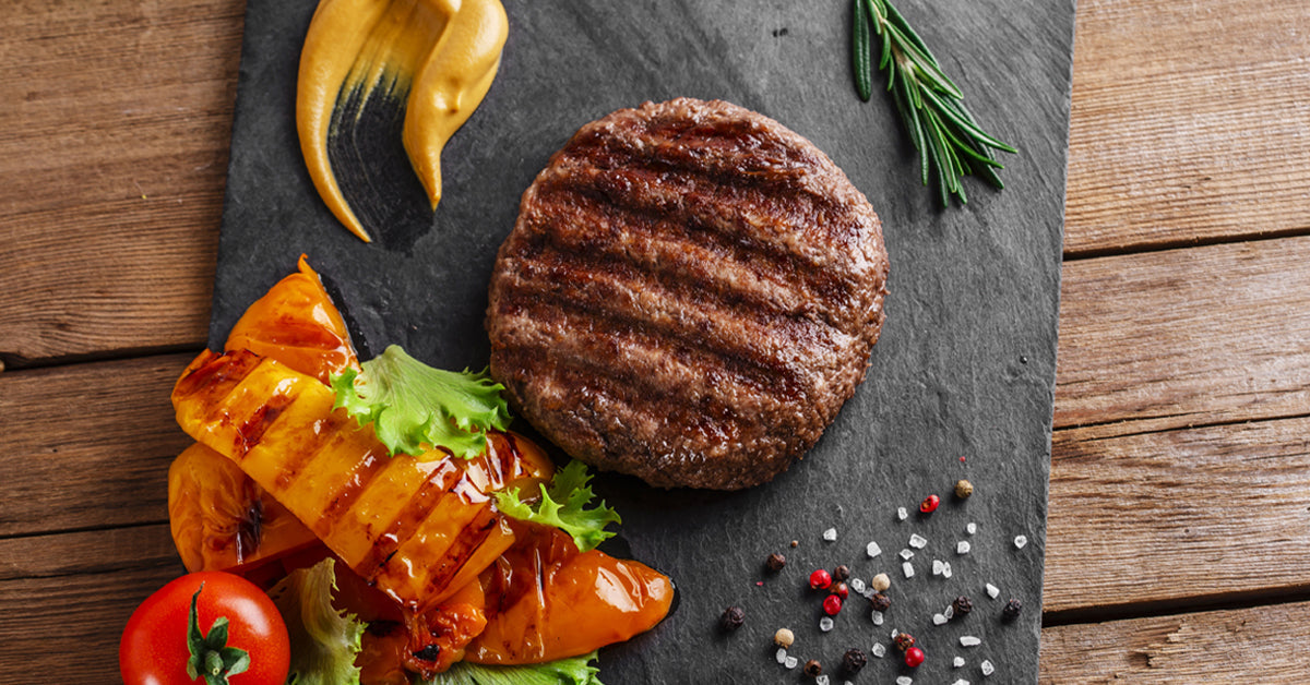 Grilled beef patty served on slate with roasted peppers, tomatoes, lettuce, mustard, and herbs.