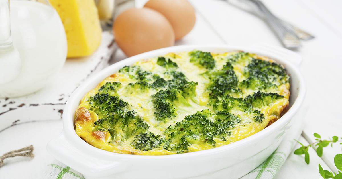Baked broccoli and cheese casserole in a white dish, surrounded by eggs, cheese, and kitchen utensils.