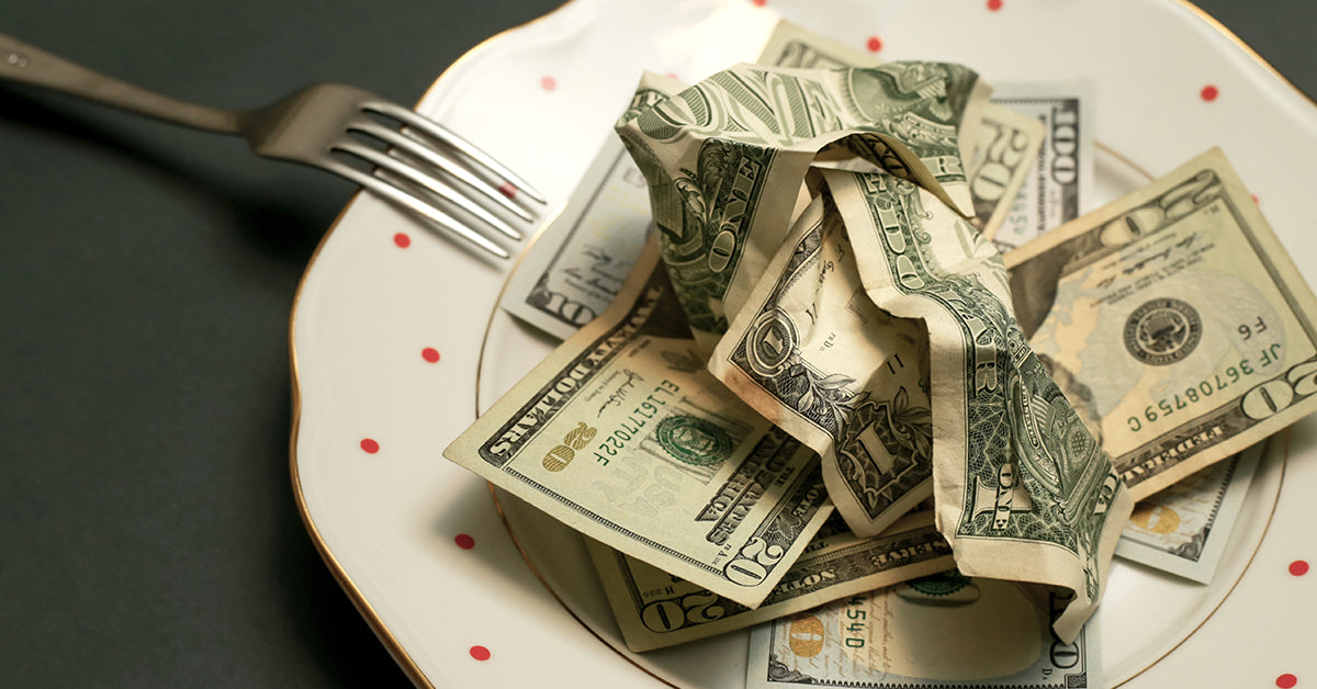 A fork rests beside a white plate with red polka dots, which is filled with crumpled U.S. dollar bills, symbolizing a commentary on money and consumption.