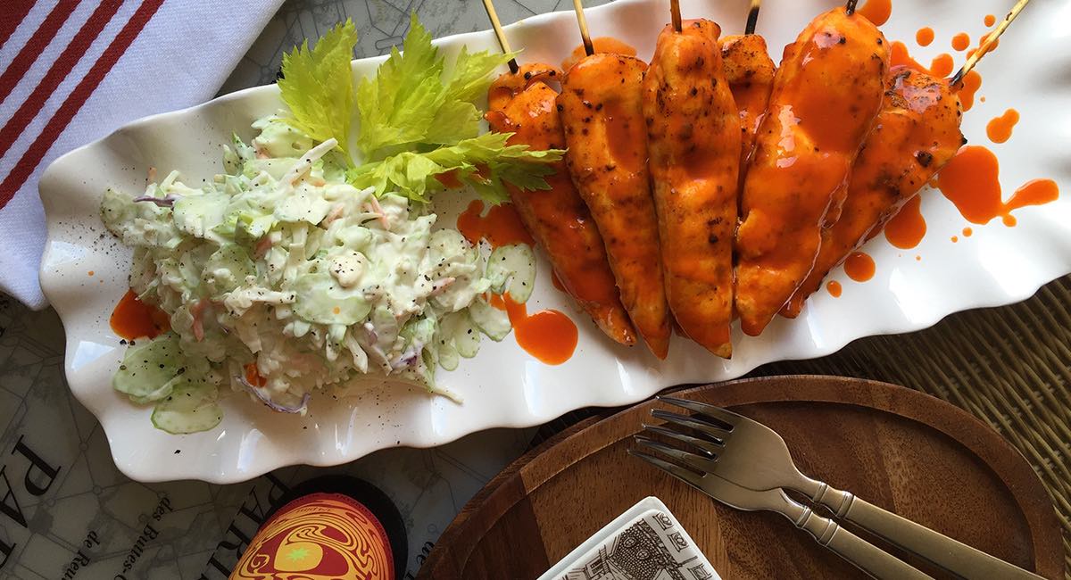 Grilled chicken skewers coated in orange sauce are served on a white platter alongside creamy coleslaw and garnished with lettuce, with a fork and knife on a wooden plate nearby.