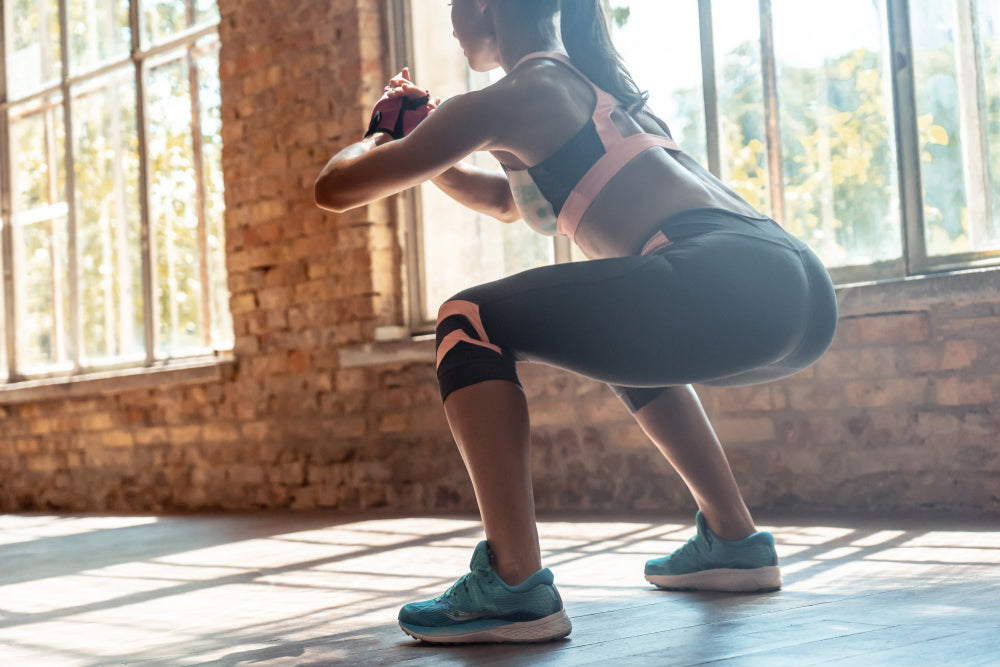 A woman in athletic wear performs a squat while holding weights in a sunlit gym with large windows and exposed brick walls, focusing on her form and strength.