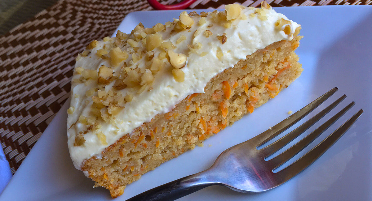 Slice of carrot cake with creamy frosting and chopped nuts on top, served on a white plate with a fork.
