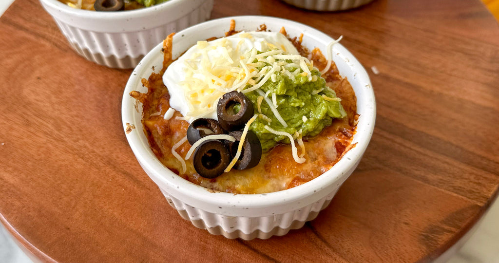 A small white ramekin filled with baked cheese topped with sour cream, guacamole, shredded cheese, and black olives sits on a wooden surface.