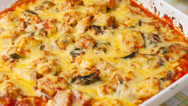 A close-up of a baked casserole featuring golden melted cheese, chunks of zucchini, and pasta, all sitting in a rich tomato sauce within a white dish.