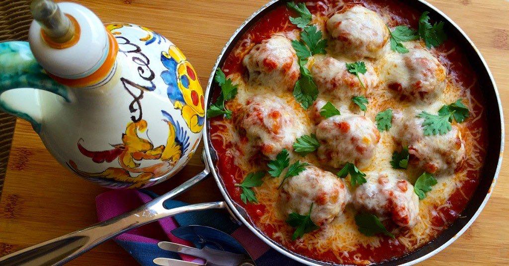 Skillet of cheesy meatballs in tomato sauce, topped with parsley, beside a colorful ceramic oil bottle.