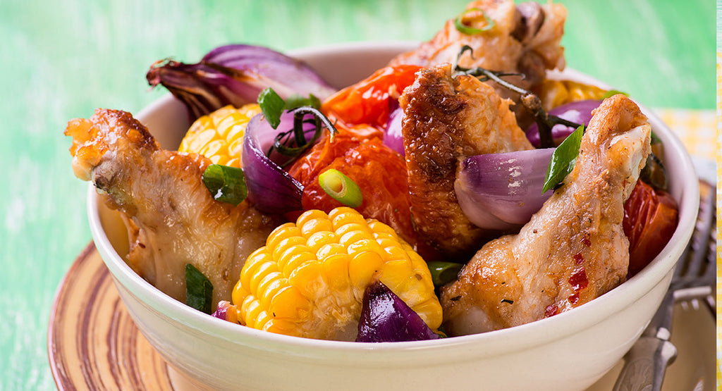 A bowl of roasted chicken wings, corn, and colorful vegetables like red onions and tomatoes is set against a green background, showcasing a vibrant and appetizing meal.
