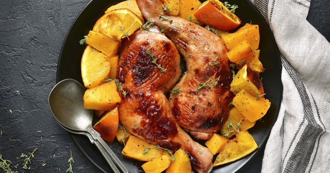 Roasted chicken legs served with seasoned pumpkin and orange pieces on a black plate with fresh herbs.