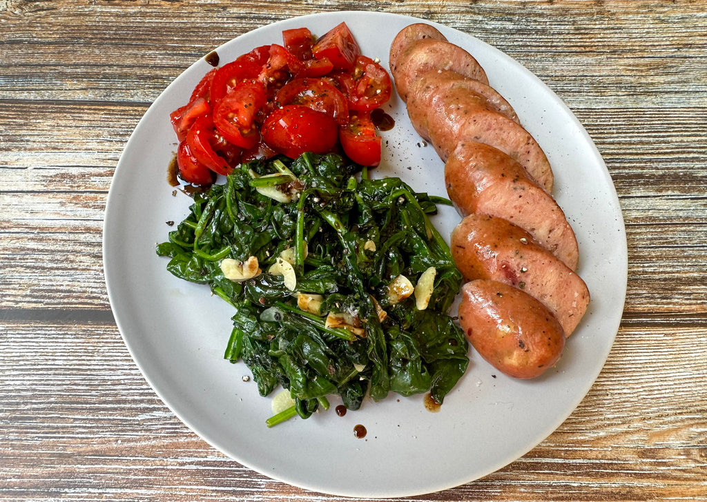 A plate features sliced sausage, sautéed spinach with garlic, and fresh diced tomatoes, all arranged on a wooden table.