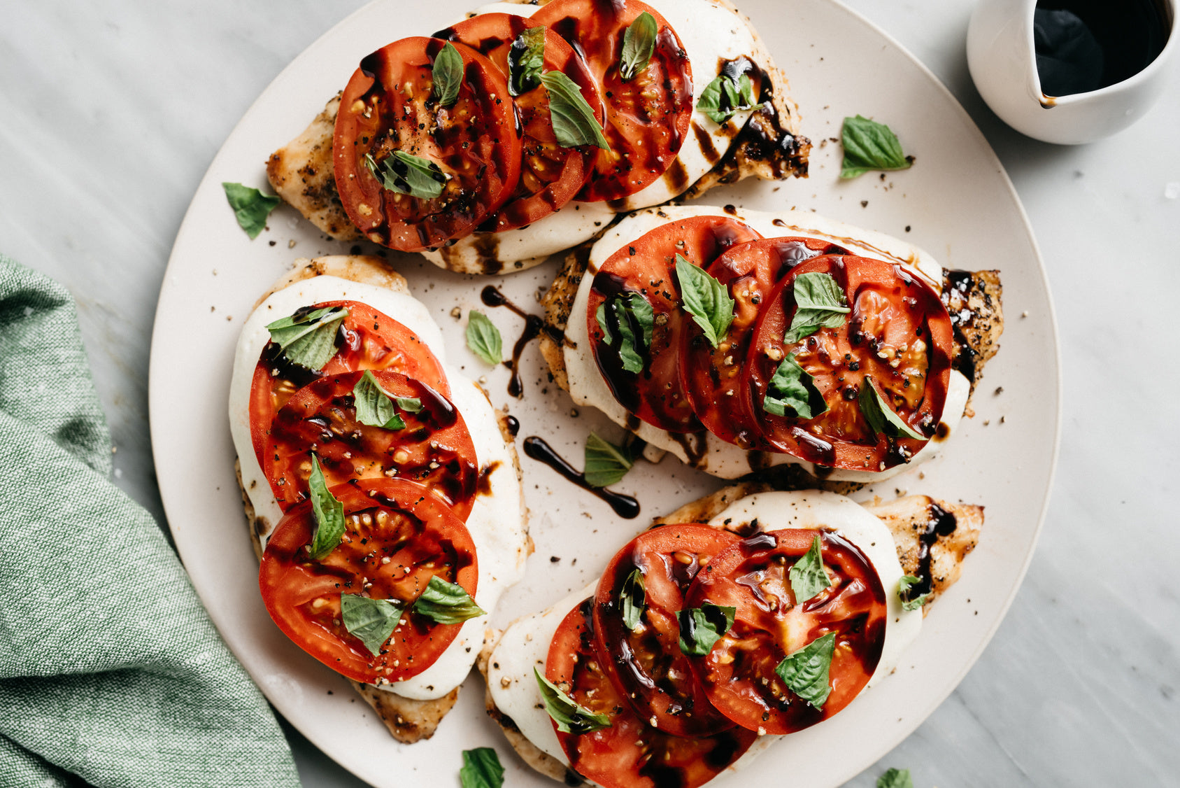 Grilled chicken breasts topped with fresh mozzarella, sliced tomatoes, and basil, drizzled with balsamic glaze, served on a round plate with a green cloth nearby.