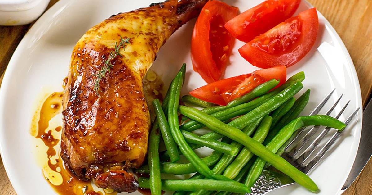 A plate features a golden-brown roasted chicken leg garnished with thyme, accompanied by green beans and sliced tomatoes, set on a wooden table with a fork beside it.