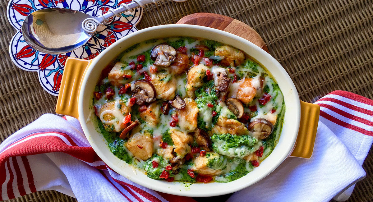 Baked chicken with spinach sauce, mushrooms, melted cheese, and red peppers in a casserole dish.