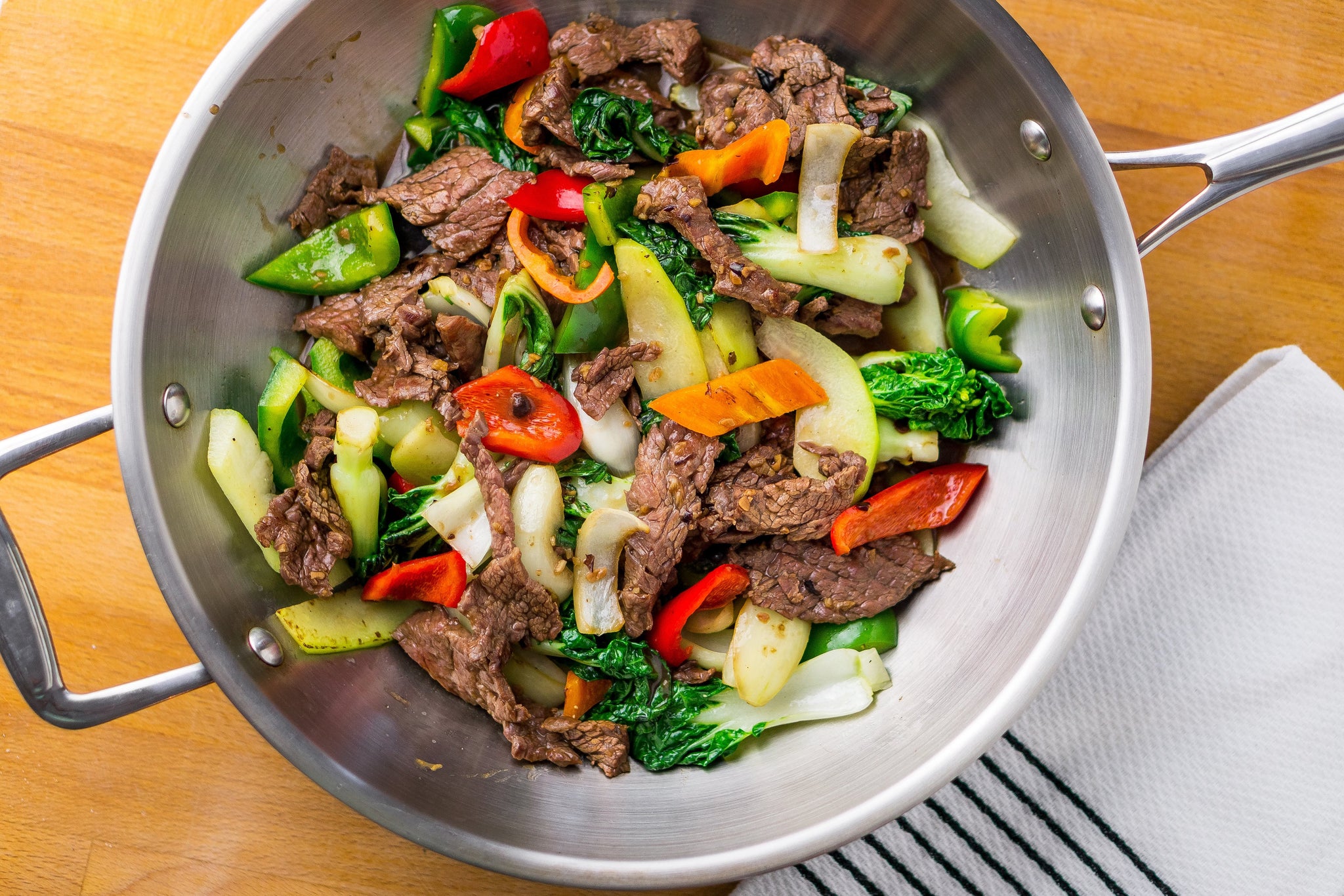 Stir-fried beef with mixed vegetables including peppers, bok choy, and zucchini in a stainless-steel pan.