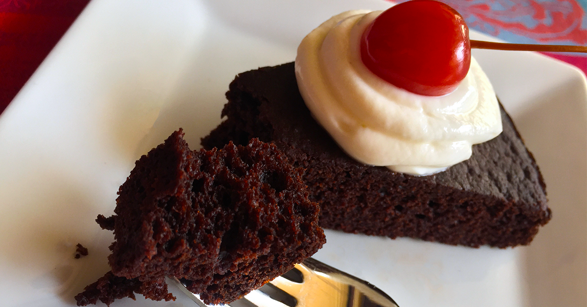 Slice of chocolate cake with whipped cream and a cherry on top, with a fork holding a bite-sized piece.