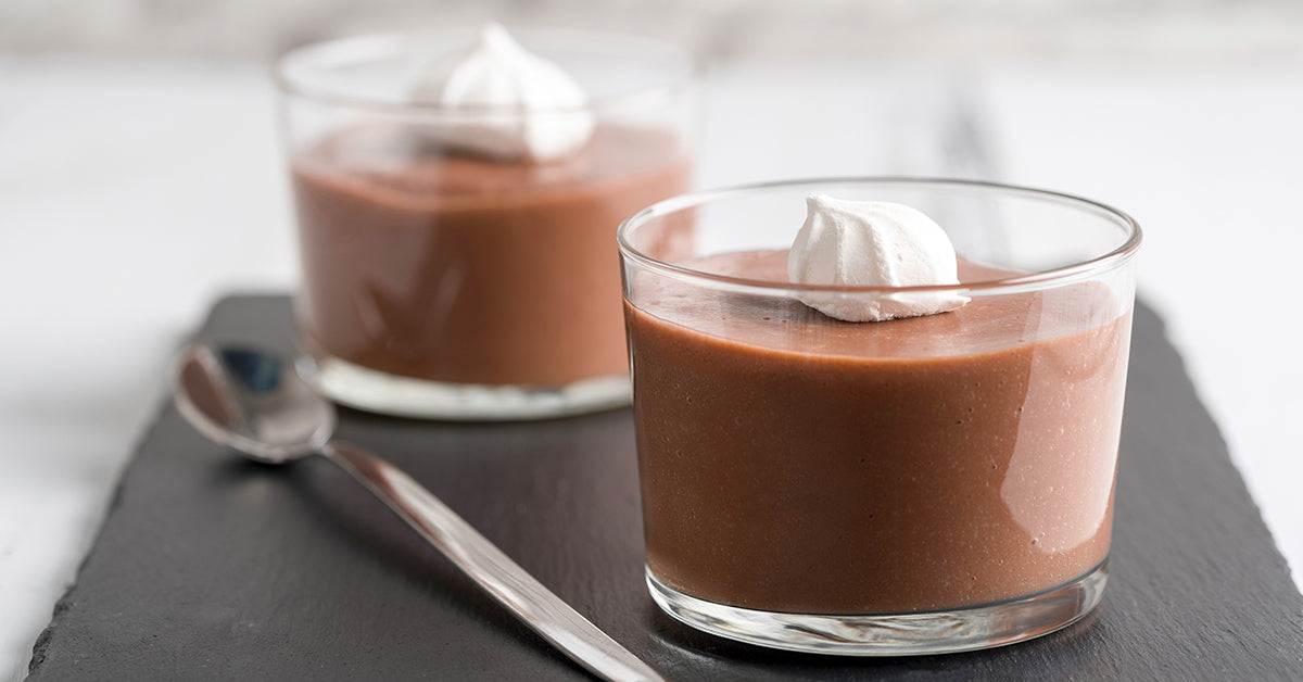 Two glasses of chocolate pudding topped with whipped cream, served on a slate board with a spoon.