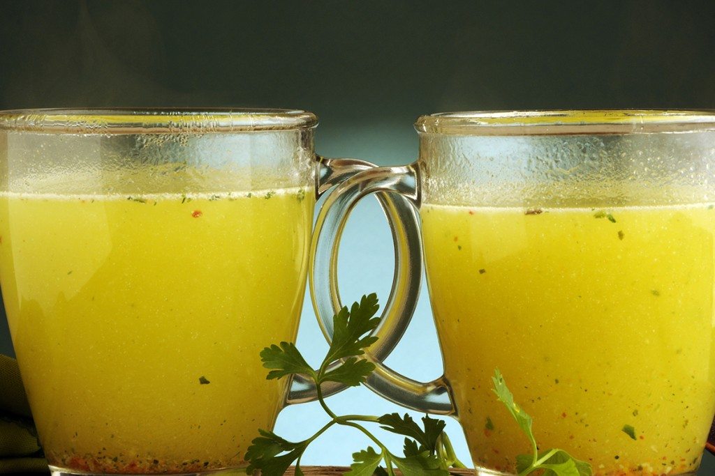 Two steaming glass mugs of yellow seasoned broth with herbs.
