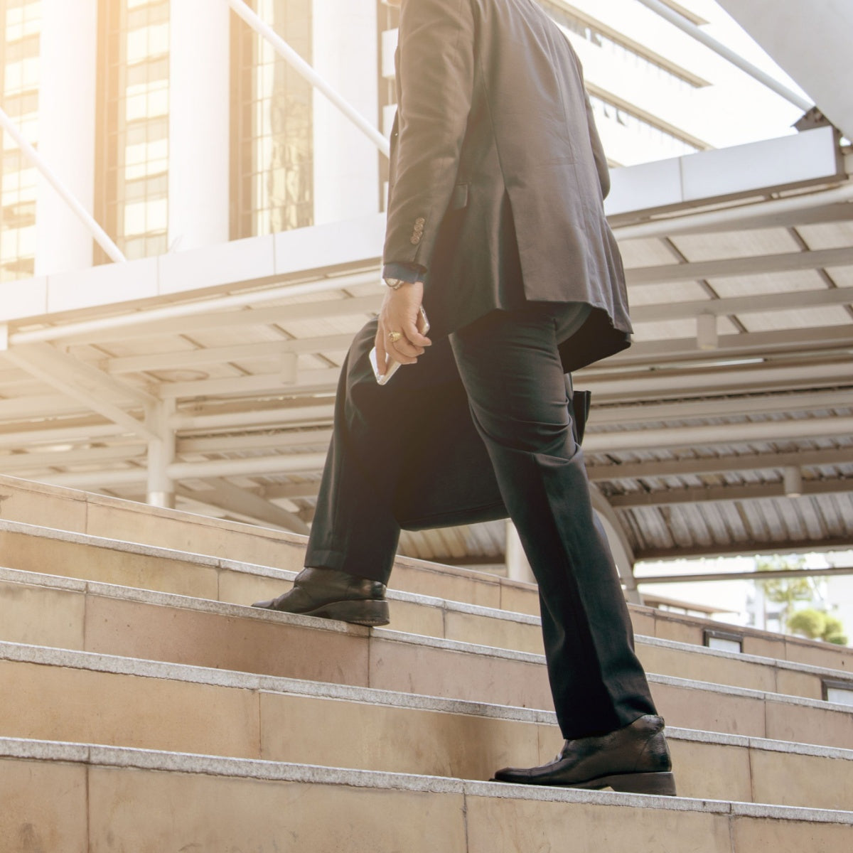 A man in a suit climbs outdoor steps while holding a phone, with a modern building and sunlight in the background, suggesting a busy urban environment.