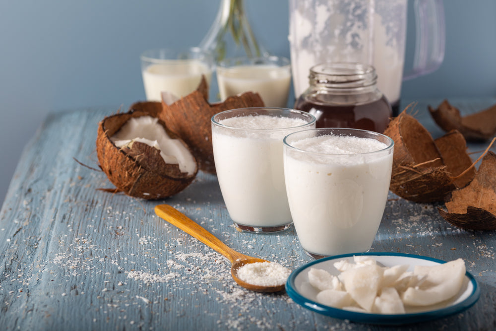 Two glasses of creamy coconut drink sit on a blue wooden table, surrounded by coconut shells, a spoon with shredded coconut, and a jar of honey, suggesting a tropical beverage preparation.